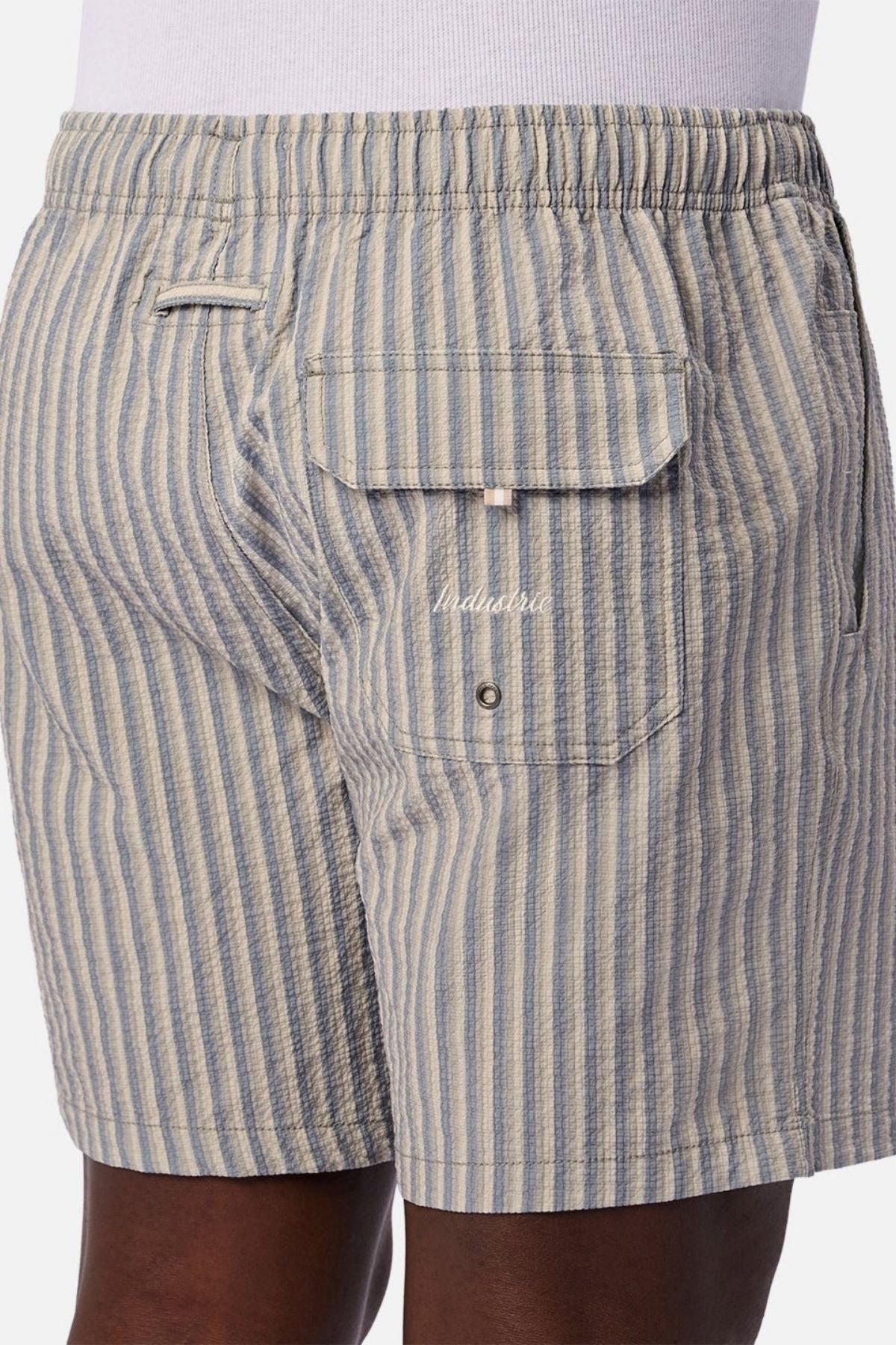 A person is shown from the back waist to mid-thigh, wearing Industrie The Formosa Swim Short in blue and beige stripes, featuring a back flap pocket and an elastic waistband.