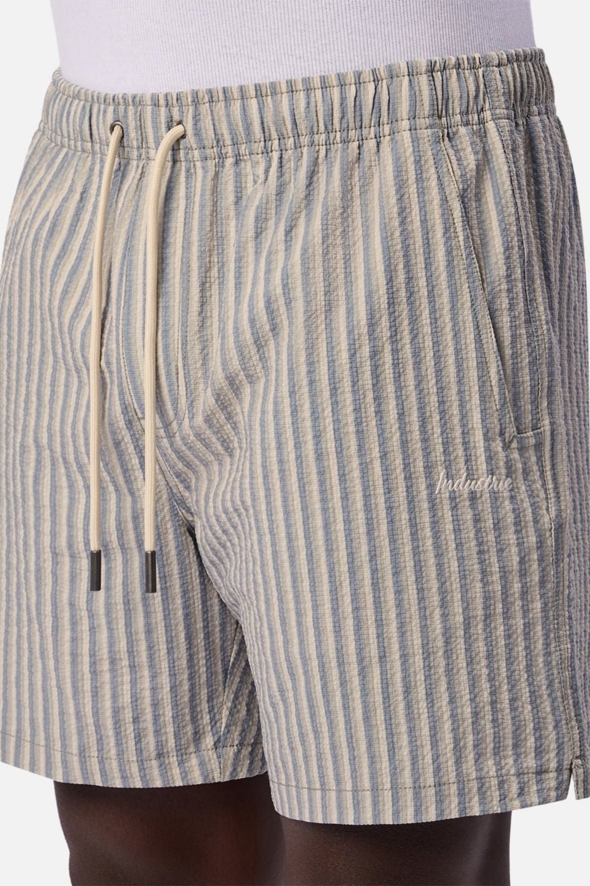 A person wears the Industrie The Formosa Swim Short in light blue and beige vertical stripes, featuring a drawstring waist, side pockets, and an embroidered logo on the left side.