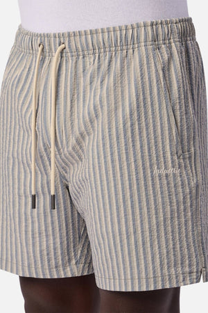 A person wears the Industrie The Formosa Swim Short in light blue and beige vertical stripes, featuring a drawstring waist, side pockets, and an embroidered logo on the left side.