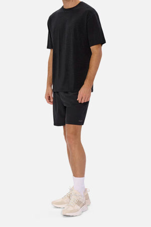 A man wears an Industrie The Freeman Athletic Tee in black, black shorts, white crew socks, and beige sneakers while standing against a plain white background. His head is not visible in the image.