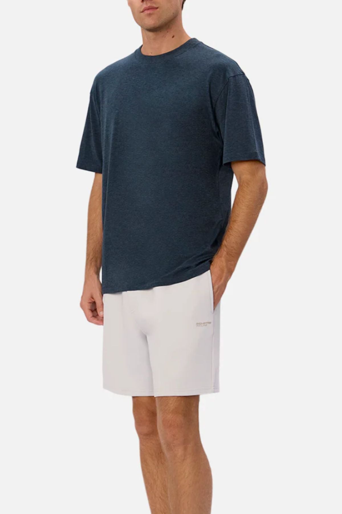 A man wears the Industrie The Freeman Athletic Tee in a relaxed, dark blue style with light shorts, standing with one hand in his pocket against a plain white background.