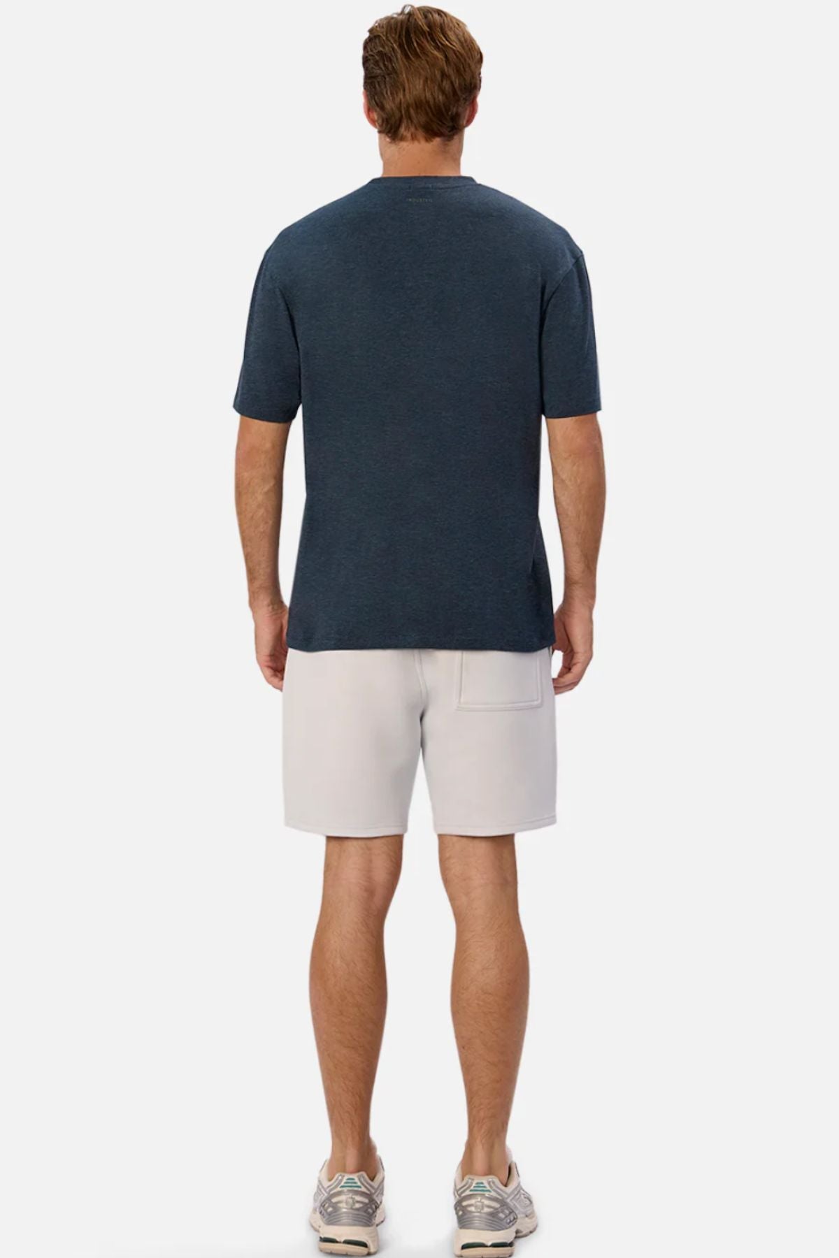 A man faces away from the camera, wearing the Industrie The Freeman Athletic Tee in dark blue, paired with light gray shorts featuring a back pocket and gray sneakers, set against a plain light background.