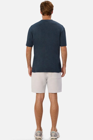 A man faces away from the camera, wearing the Industrie The Freeman Athletic Tee in dark blue, paired with light gray shorts featuring a back pocket and gray sneakers, set against a plain light background.