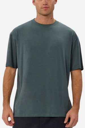 A man wearing the Industrie The Freeman Athletic Tee—a relaxed fit, short-sleeve, dark green lightweight t-shirt—stands facing forward against a plain, light background. His face is partially visible.