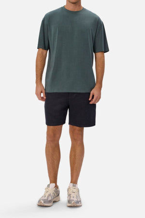 A person dressed in a dark green Industrie The Freeman Athletic Tee, black athletic shorts, and light-colored sneakers stands against a plain, light background. Their hands rest relaxed by their sides, obscuring any visible branding. The individual's face remains out of view.