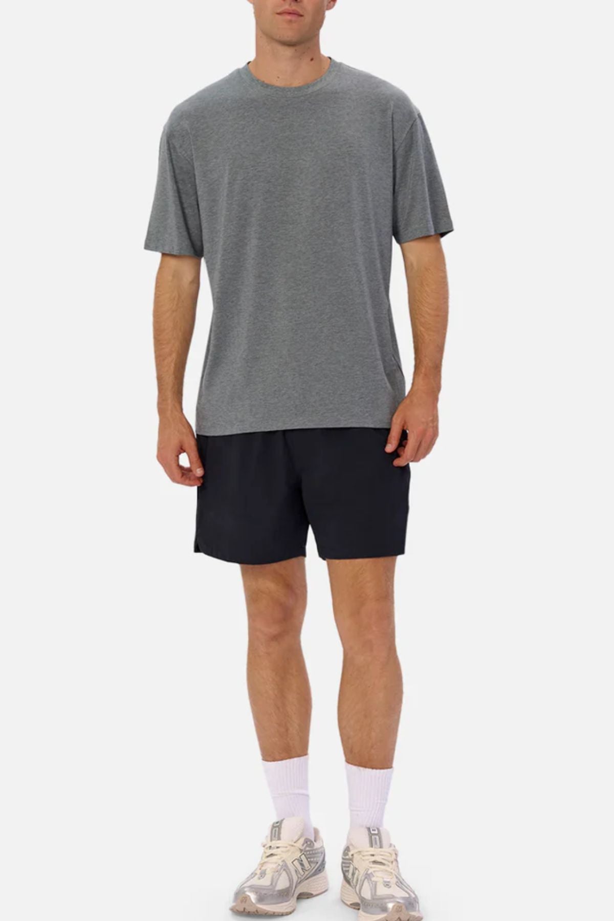 A man stands against a plain background wearing the Industrie The Freeman Athletic Tee in grey, paired with black shorts, white socks, and white sneakers.