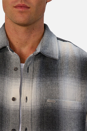 A man wears the Industrie The Highland Shirt in a tailored fit, showcasing its black, white, and gray shadow check pattern over a white T-shirt. The shirt features a small chest pocket and visible buttons from neck to mid-chest.