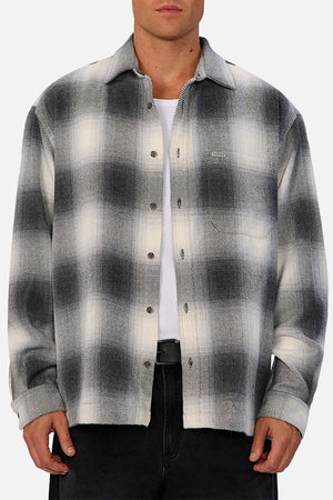 A person wears the Industrie The Highland Shirt in black and white shadow check, untucked with the top button undone, over a white T-shirt and black pants, standing against a light gray background, highlighting its tailored fit.