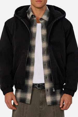 A person wears the Industrie The Illinois Jacket, a black zip-up hooded jacket, over a plaid shirt and white tee with olive green pants for a casual, layered style perfect for cooler weather.
