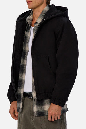 Wearing the Industrie The Illinois Jacket—a black hooded, heavyweight jacket—over a black-and-white plaid shirt and white tee with olive green pants, a person stands against a plain light background.
