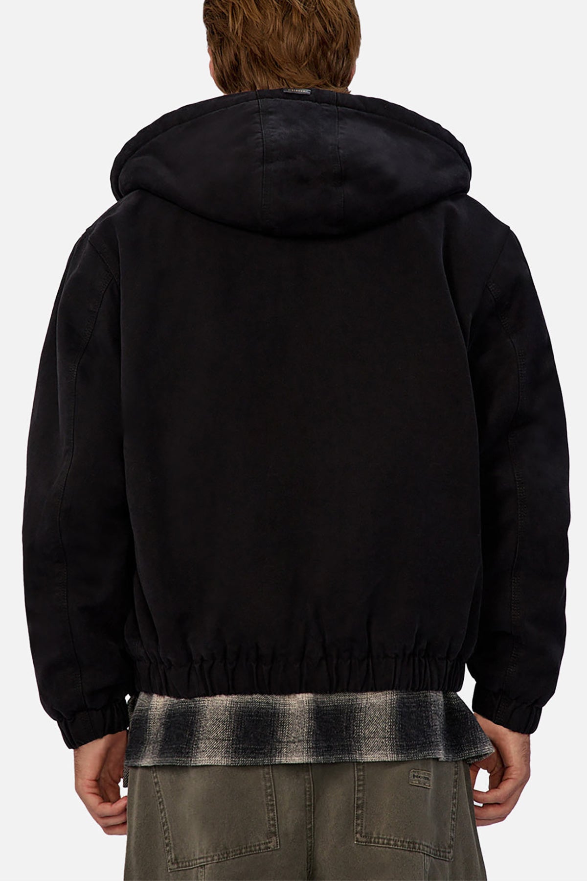 Seen from behind against a plain light background, a person with light brown hair wears the Industrie The Illinois Jacket, a black heavyweight hooded jacket, layered over a plaid shirt and paired with olive green pants.