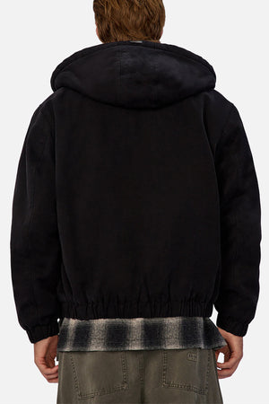 Seen from behind against a plain light background, a person with light brown hair wears the Industrie The Illinois Jacket, a black heavyweight hooded jacket, layered over a plaid shirt and paired with olive green pants.