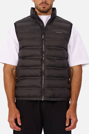 A man wears the Industrie The Kyoto Puffer Vest in black, featuring a heat-sealed design, front zipper, and subtle left-chest branding, over a white short-sleeve tee and black pants, standing against a plain light background.