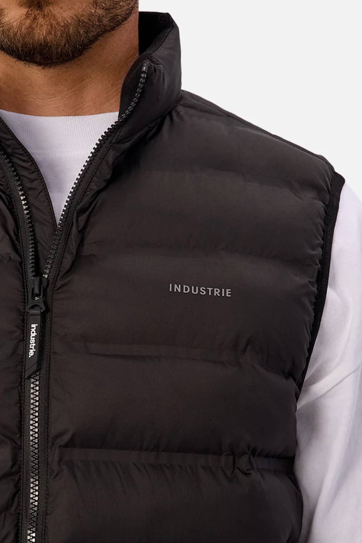 Close-up of a person wearing the Industrie The Kyoto Puffer Vest, featuring a black heat-sealed design with "INDUSTRIE" on the chest, layered over a white shirt. Only the lower face and upper torso are visible.