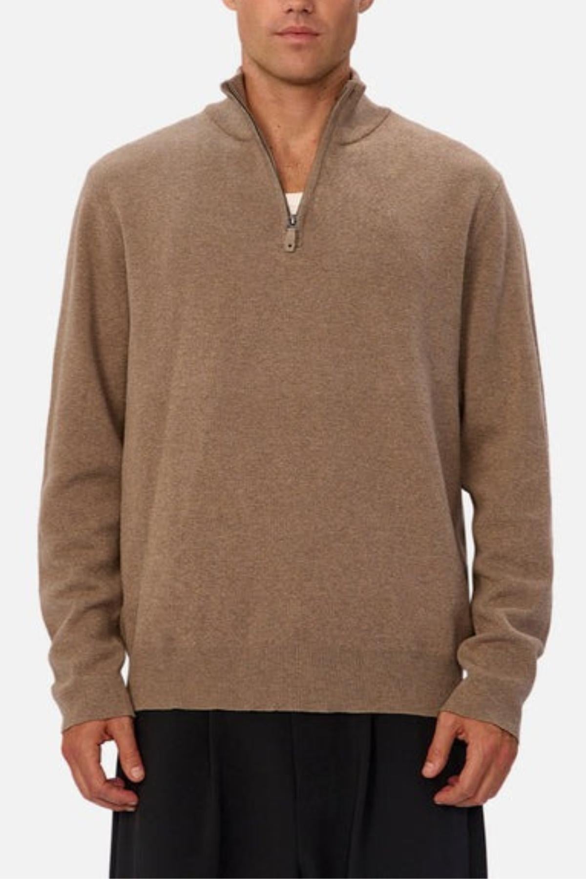 A person wears the Industrie The Lakewood Zip Neck Knit—a light brown cotton jumper with a small collar, relaxed fit, and rib knit cuffs—over a white undershirt and loose dark pants, standing against a plain light background.