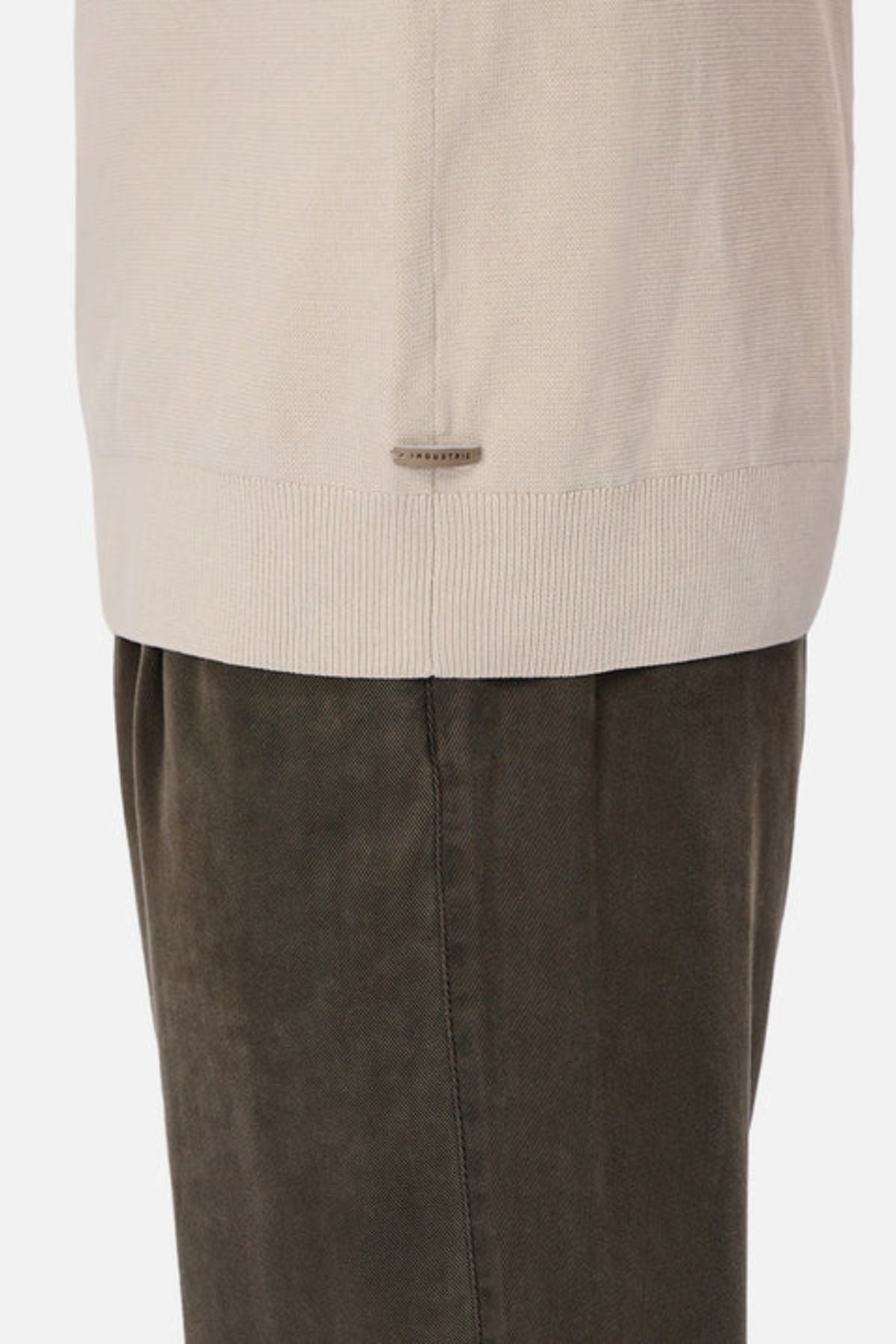A close-up of a person wearing dark olive green pants and the Industrie The Lakewood Zip Neck, a beige cotton jumper with ribbed hem, rib knit cuffs, and a small metallic tag near the bottom edge.