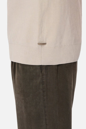 A close-up of a person wearing dark olive green pants and the Industrie The Lakewood Zip Neck, a beige cotton jumper with ribbed hem, rib knit cuffs, and a small metallic tag near the bottom edge.