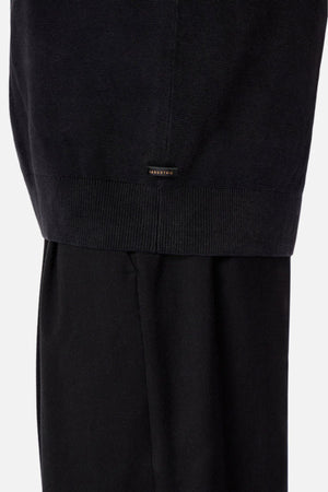 Close-up of the lower part of a person wearing Industrie The Lakewood Zip Neck and black pants, highlighting the ribbed hem and a small rectangular label above it against a plain, light-colored background.