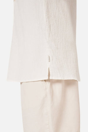 A close-up of a person wearing the Industrie The Magna Short Sleeve Shirt, an off-white textured and breathable shirt paired with cream-colored pants, highlighting the side seam and small slit at the hem that enhances its oversized fit.