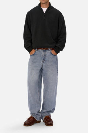A person in a black Industrie The Maine Zip Neck Knit stands against a plain background, paired with light blue baggy jeans and brown boat shoes, hands in pockets.