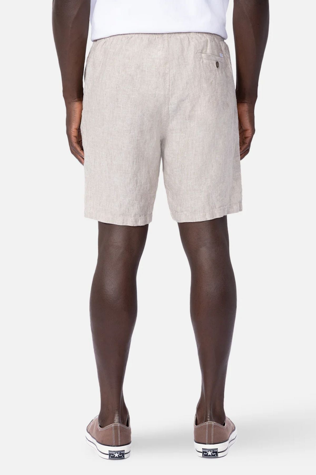 A man stands with his back to the camera in a white shirt, taupe slip-on sneakers, and Industrie The Marina Linen Short—light beige shorts celebrated for their relaxed, breathable fit. The background is plain and light gray.