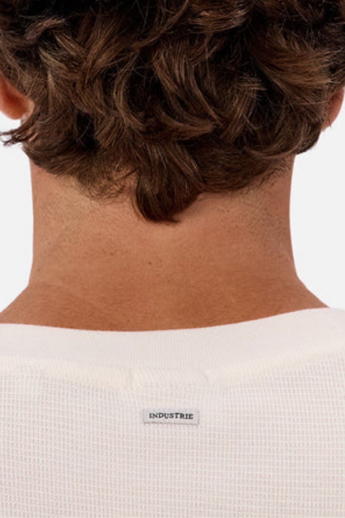 Close-up of a person's neck and shoulders wearing the Industrie The Marlon Long Sleeve Tee in white textured fabric with a small "INDUSTRIE" label. Person has short, wavy brown hair.