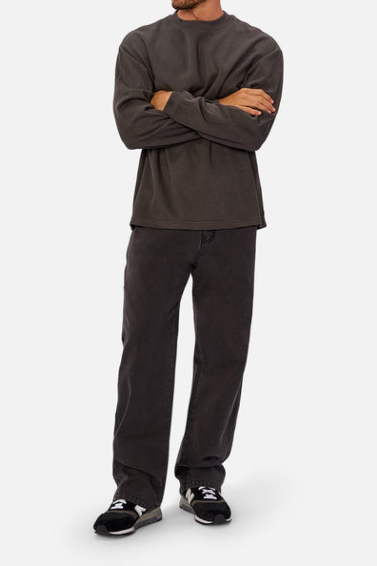 A person with arms crossed wears a loose dark gray Industrie The Marlon Long Sleeve Tee (100% cotton), matching pants, and black sneakers with white soles. Head not visible. Background is plain light gray.