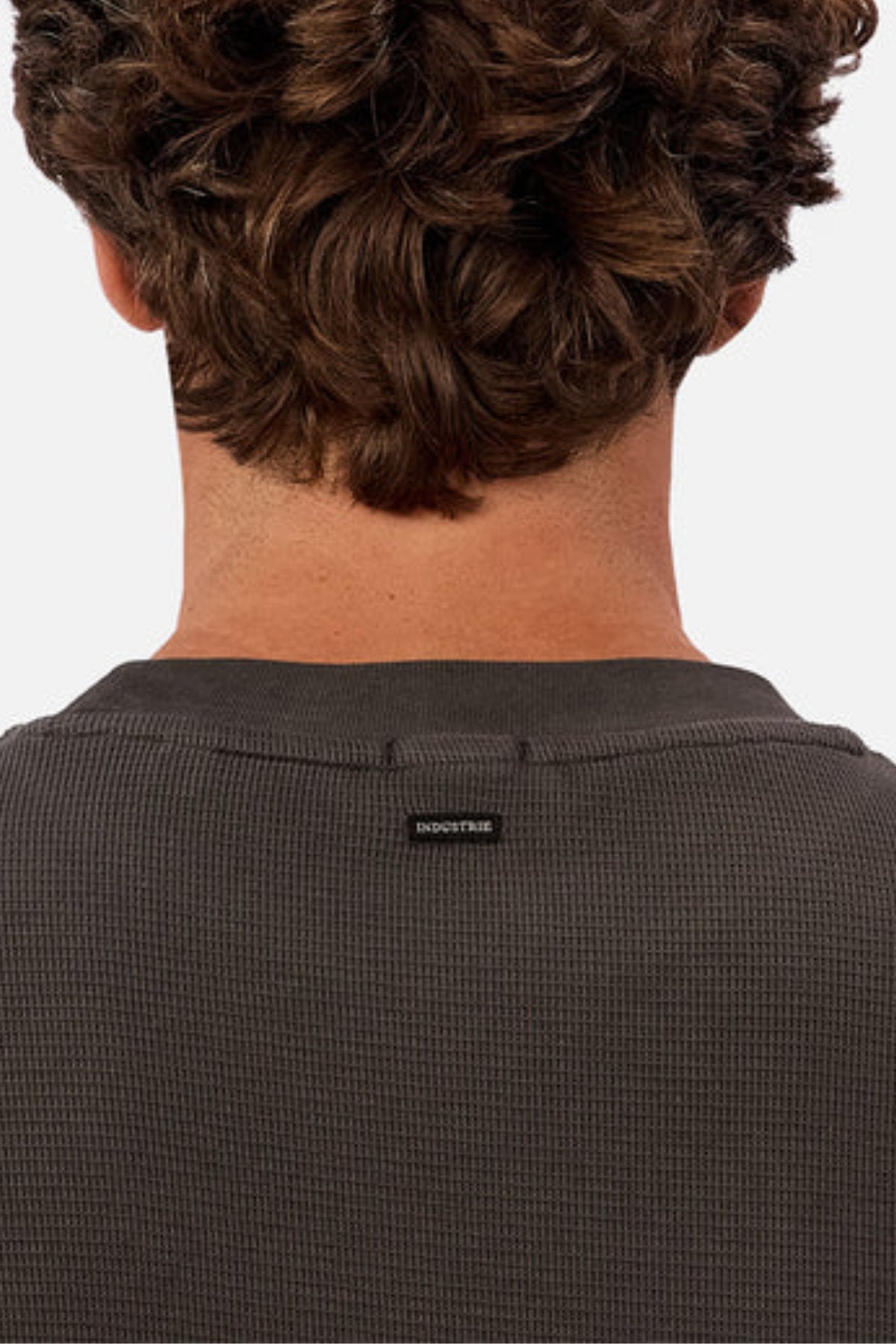 Close-up of the back of a person with curly brown hair, wearing the Industrie The Marlon Long Sleeve Tee in textured dark fabric, featuring a small black “INDUSTRIE” label on the upper back.