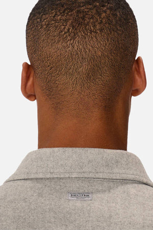 Close-up of the back of a person's head and neck wearing a light grey, relaxed-fit collared shirt. A small rectangular tag at the top center evokes the classic collar style of Industrie The New Coleman Jacket.