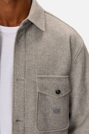A person wears the Industrie The New Coleman Jacket—a light gray, relaxed fit wool shirt jacket with a classic collar—over a white t-shirt. It features a chest flap pocket with button and an "INDUSTRIE" logo tag.