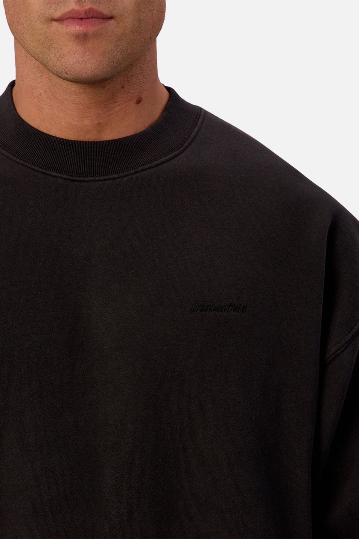 A close-up shows a person in the Industrie The New Del Sur Sweatshirt, an oversized black crewneck with subtle embroidery on the left chest, showcasing premium construction and branding against a light background.