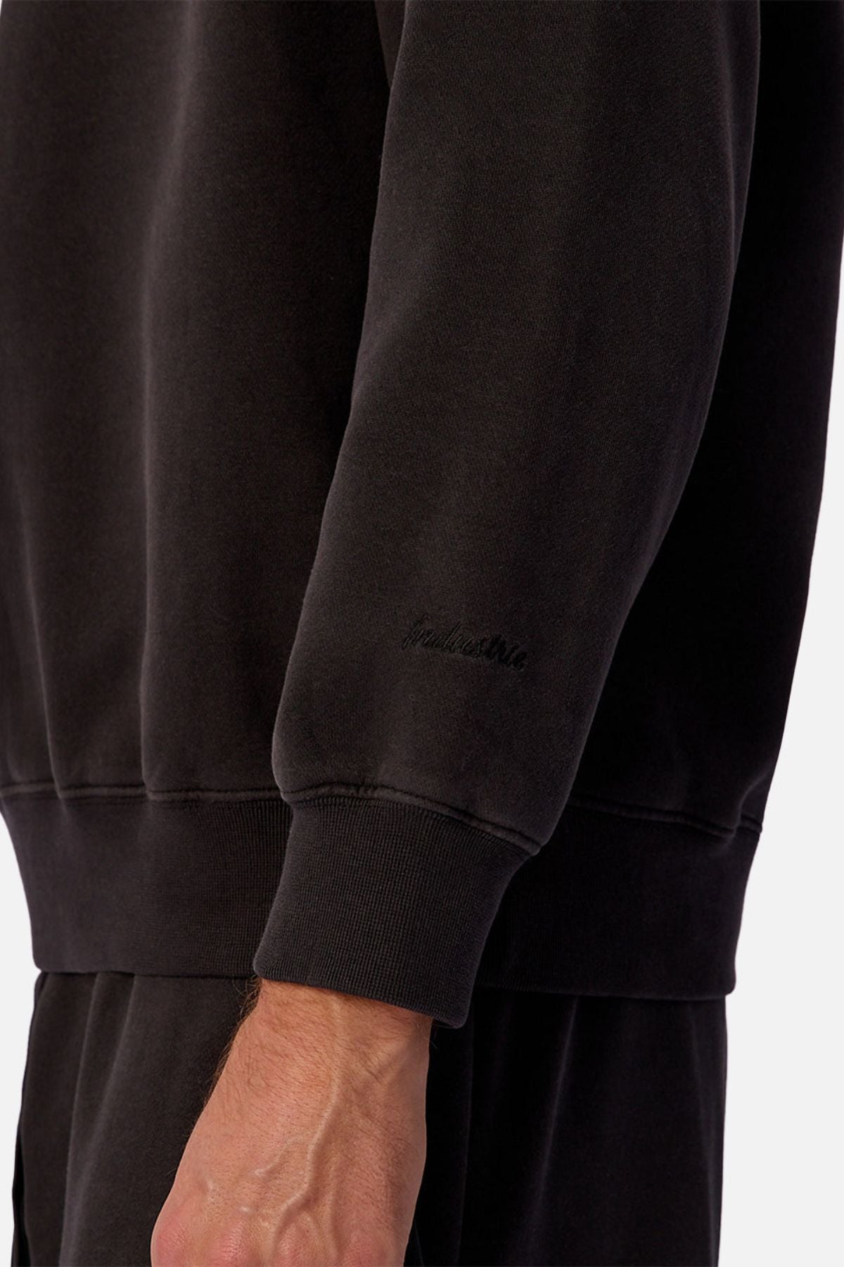 A close-up of a person wearing the Industrie The New Del Sur Sweatshirt in black with matching pants. Subtle embroidered text appears near the wrist, showcasing premium construction. The background is plain and light-colored.