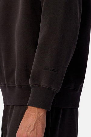 A close-up of a person wearing the Industrie The New Del Sur Sweatshirt in black with matching pants. Subtle embroidered text appears near the wrist, showcasing premium construction. The background is plain and light-colored.