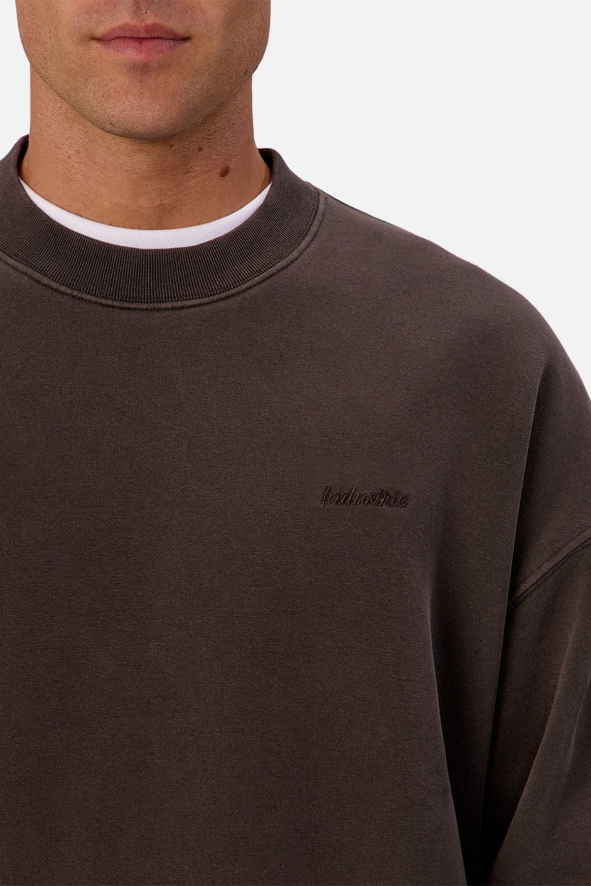 A close-up of a person wearing the dark brown Industrie The New Del Sur Sweatshirt, layered over a white t-shirt. The embroidered logo is visible on the left chest, highlighting its premium construction. Only their chin and lower face are shown.