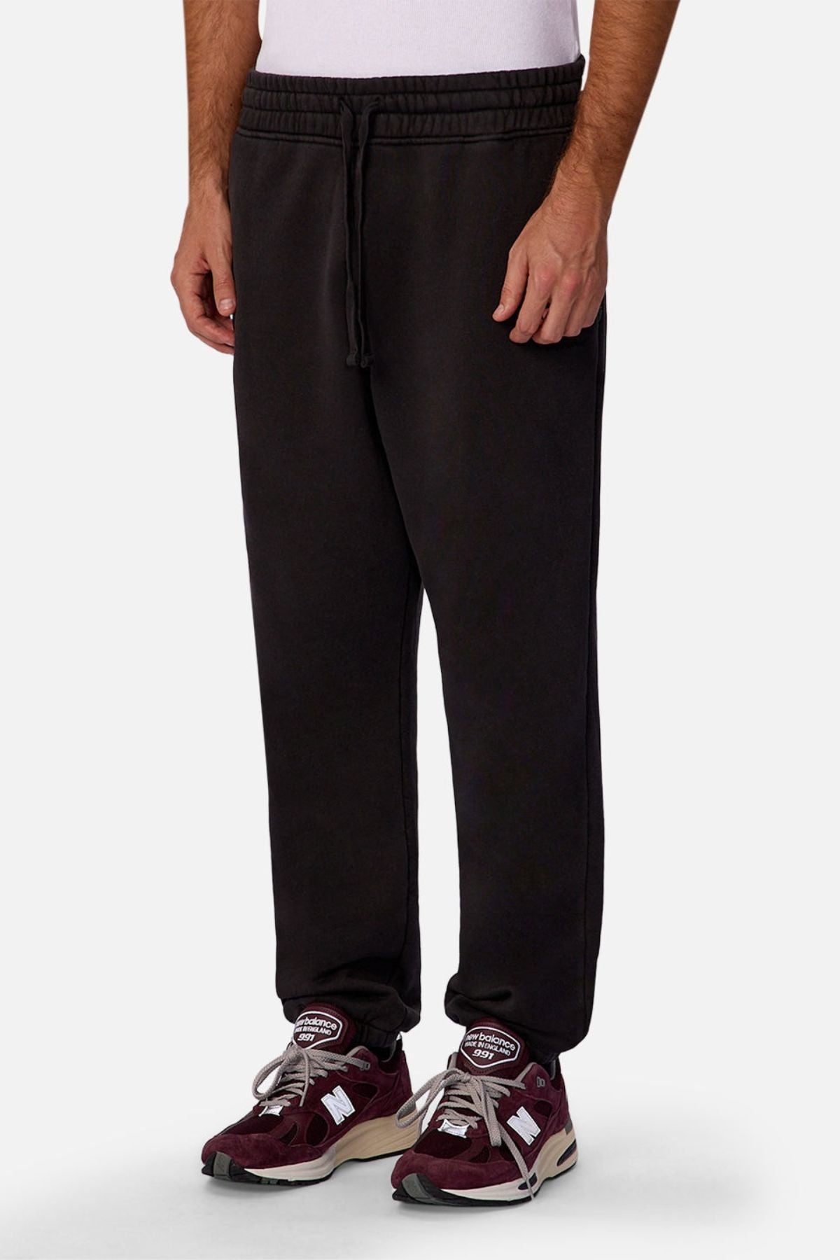 A person wearing Industrie The New Del Sur Trackpant in black, a white shirt, and maroon New Balance sneakers stands against a plain light gray background.