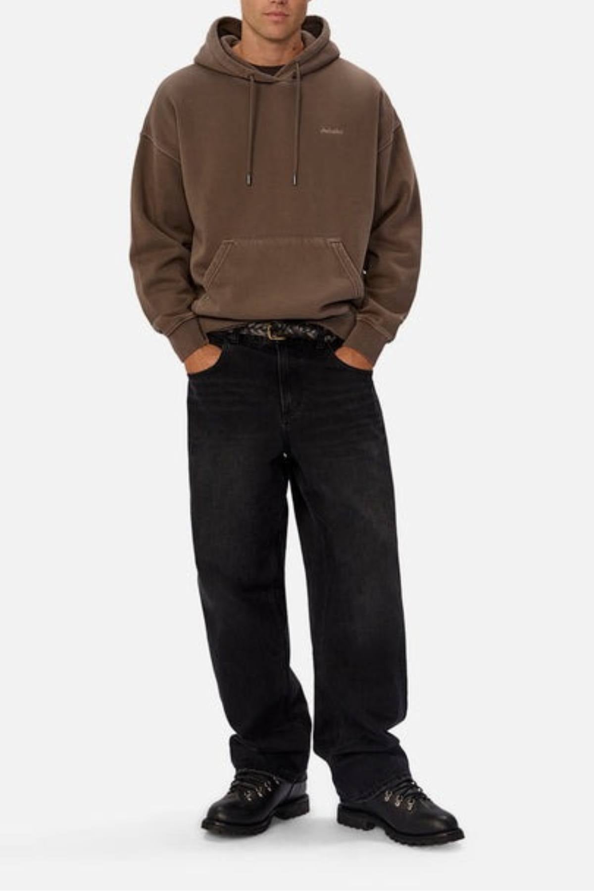 A person stands with hands in the pockets of black jeans, wearing the Industrie The New Del Sur Washed Hoodie in brown and black shoes, against a plain light-colored background.