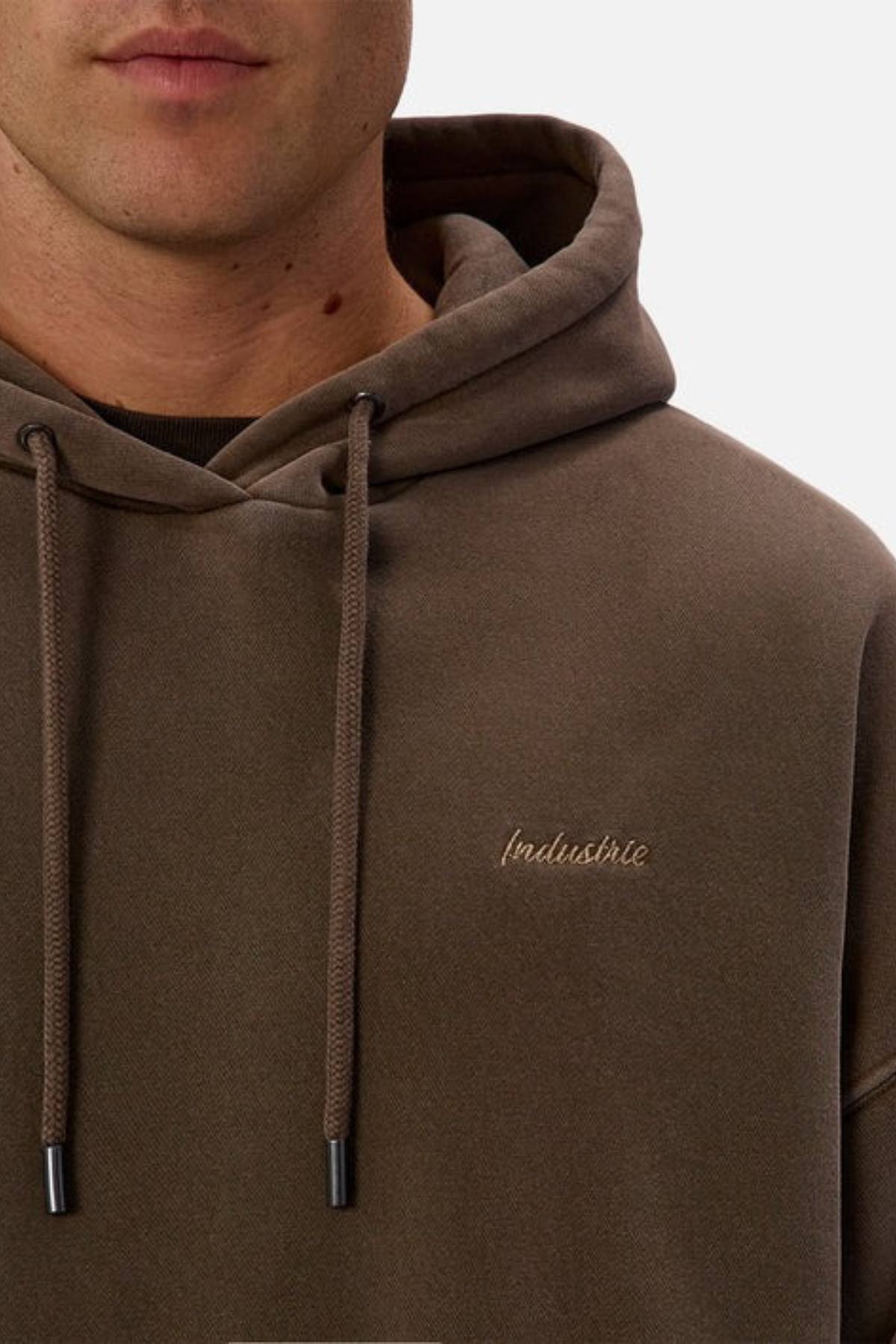 A close-up of a person wearing the Industrie The New Del Sur Washed Hoodie in brown acid wash, featuring drawstrings, "Industrie" chest embroidery, and an oversized fit for a relaxed look. The person’s face is partially visible.