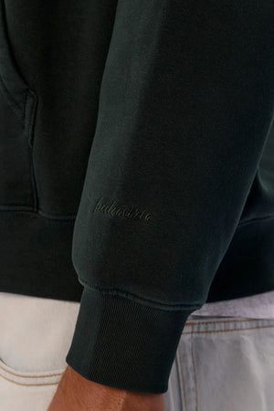 A close-up of the Industrie The New Del Sur Washed Hoodie sleeve in dark green, featuring "inclusive" embroidery in matching thread, shown beside light-colored pants with visible stitching.