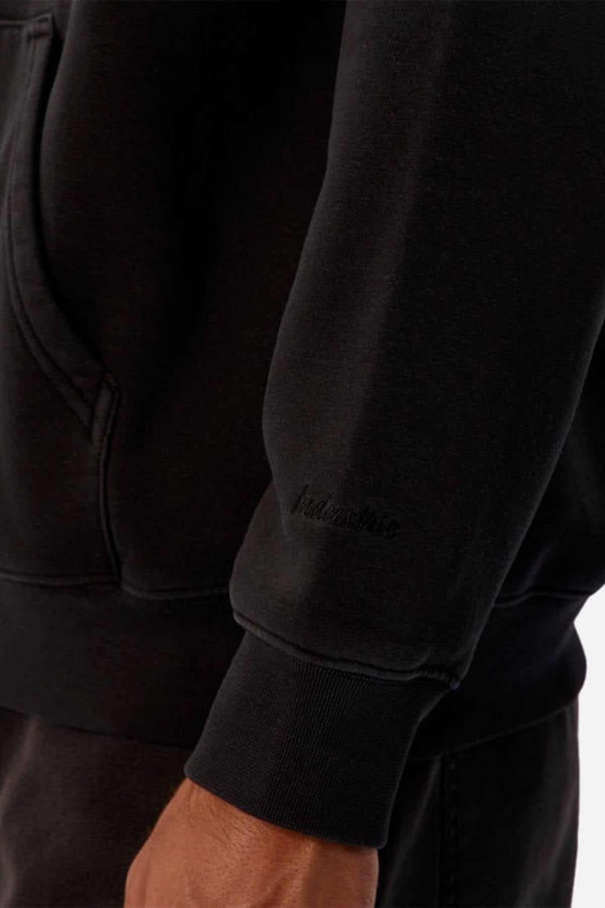 A close-up shows someone wearing the Industrie The New Del Sur Washed Hoodie in black, featuring subtle embroidered text on the acid wash sleeve. Their arm and hand are visible along with a front pocket seam, against a neutral background.