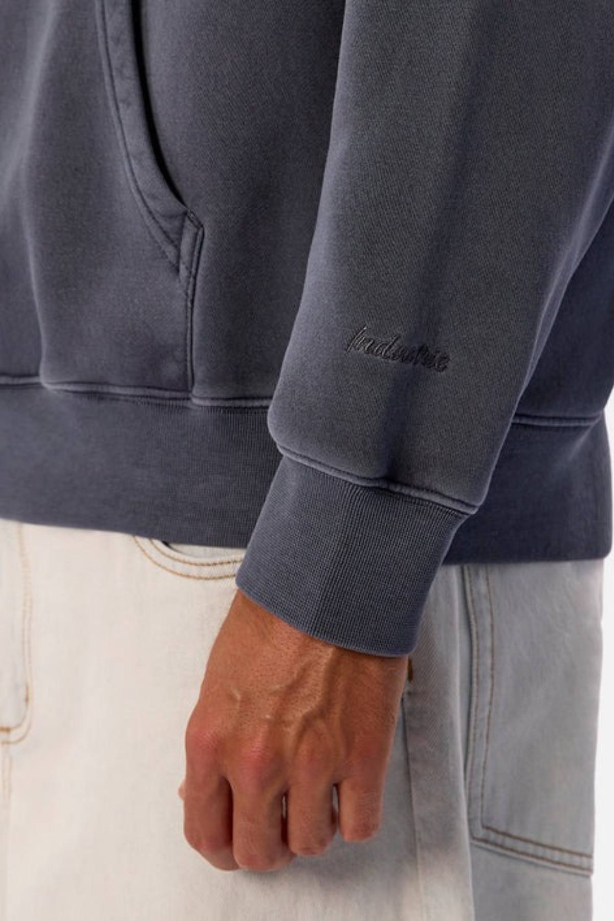 A person wearing the Industrie The New Del Sur Washed Hoodie in an oversized faded dark gray style with sleeve embroidery, paired with light-colored jeans and their hand resting by their side.