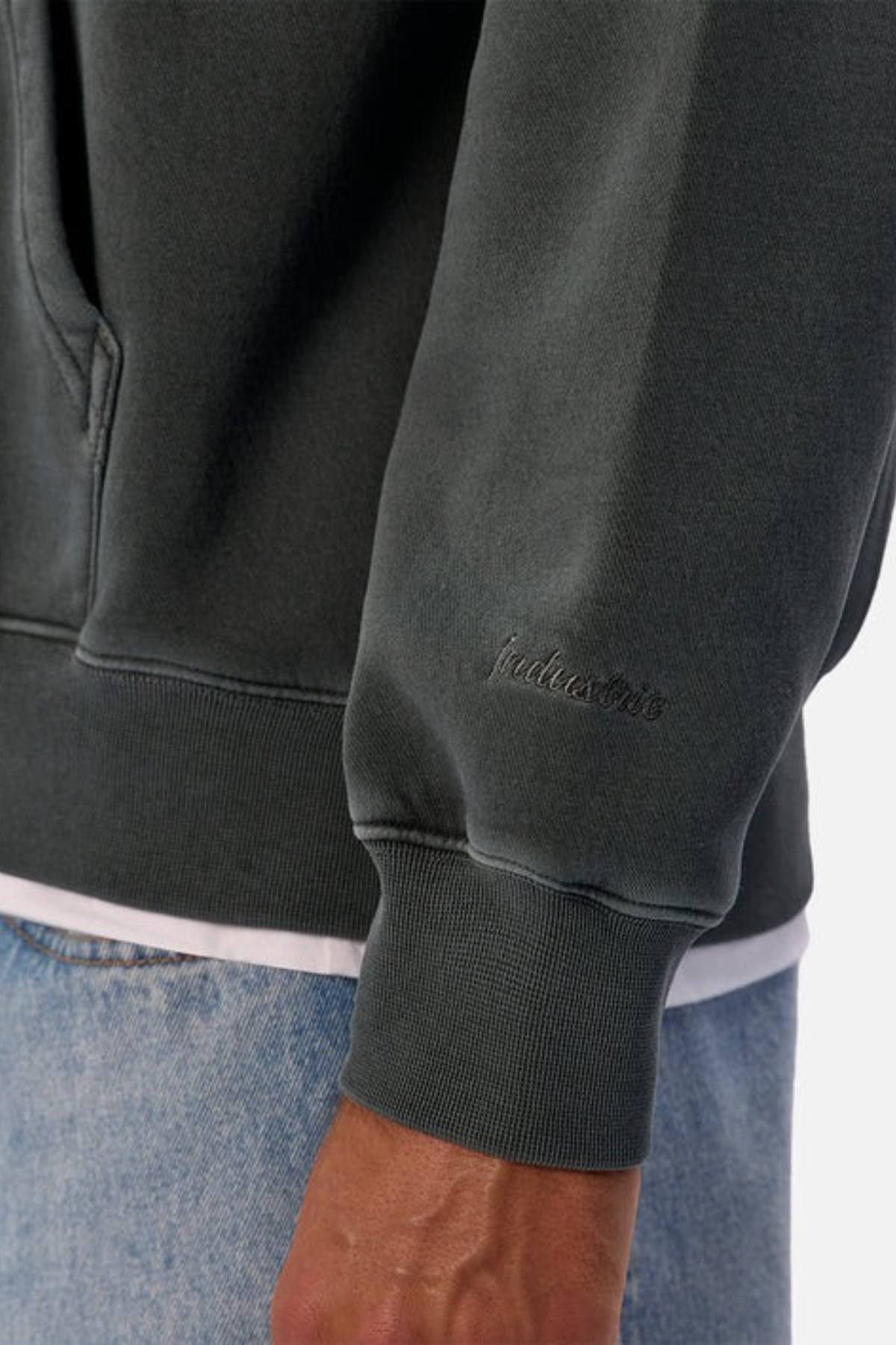 A close-up of a person wearing the Industrie The New Del Sur Washed Hoodie in dark gray with "hallows" sleeve embroidery, light blue jeans, and a white shirt underneath peeking out.