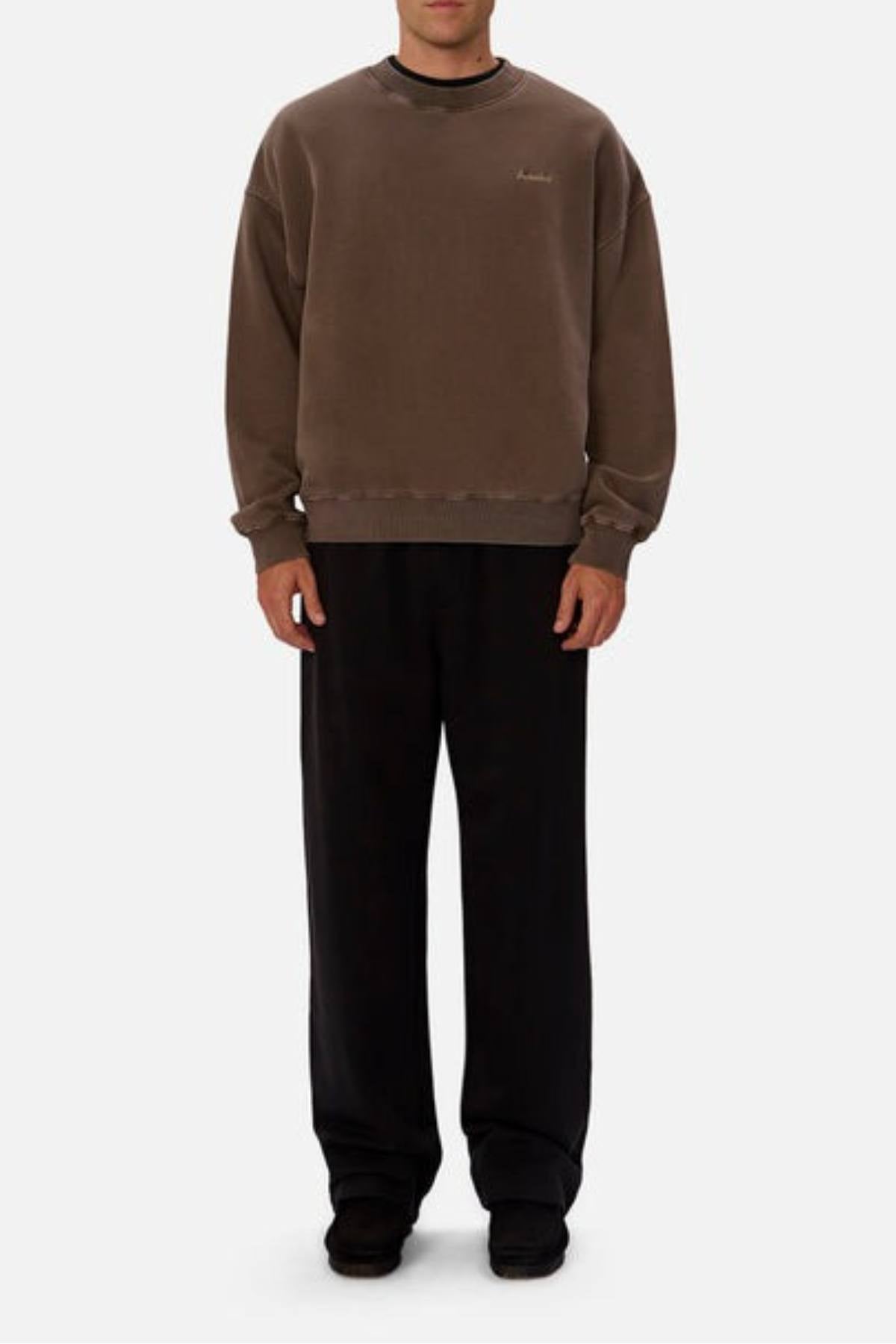 A person stands against a plain background, wearing an oversized Industrie The New Del Sur Washed Sweat and wide-leg black pants with black shoes. Their hands are by their sides and their face is partially visible.