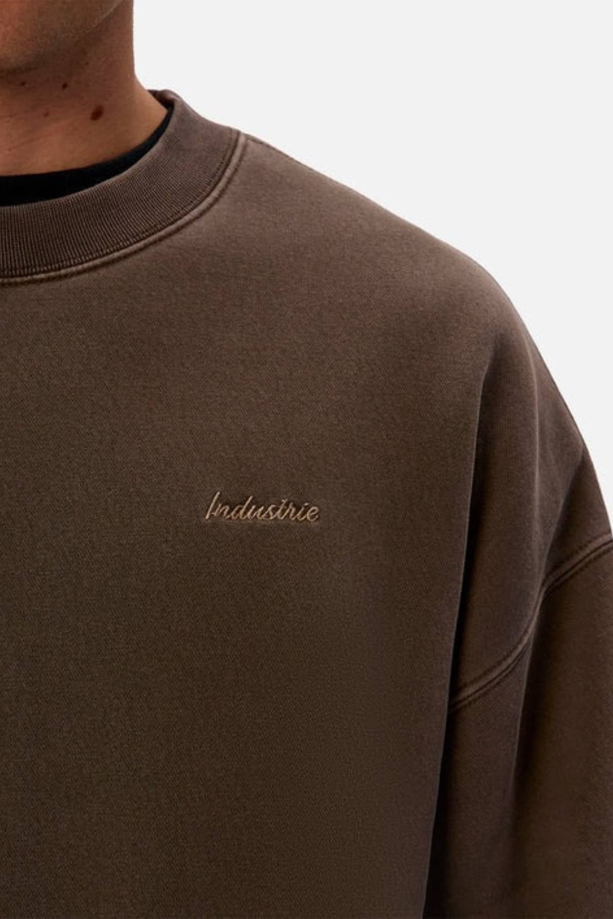 A close-up shows a person in an oversized fit brown Industrie The New Del Sur Washed Sweat, with the Industrie logo embroidered in small, light letters on the chest. Only their lower face and upper torso are visible.