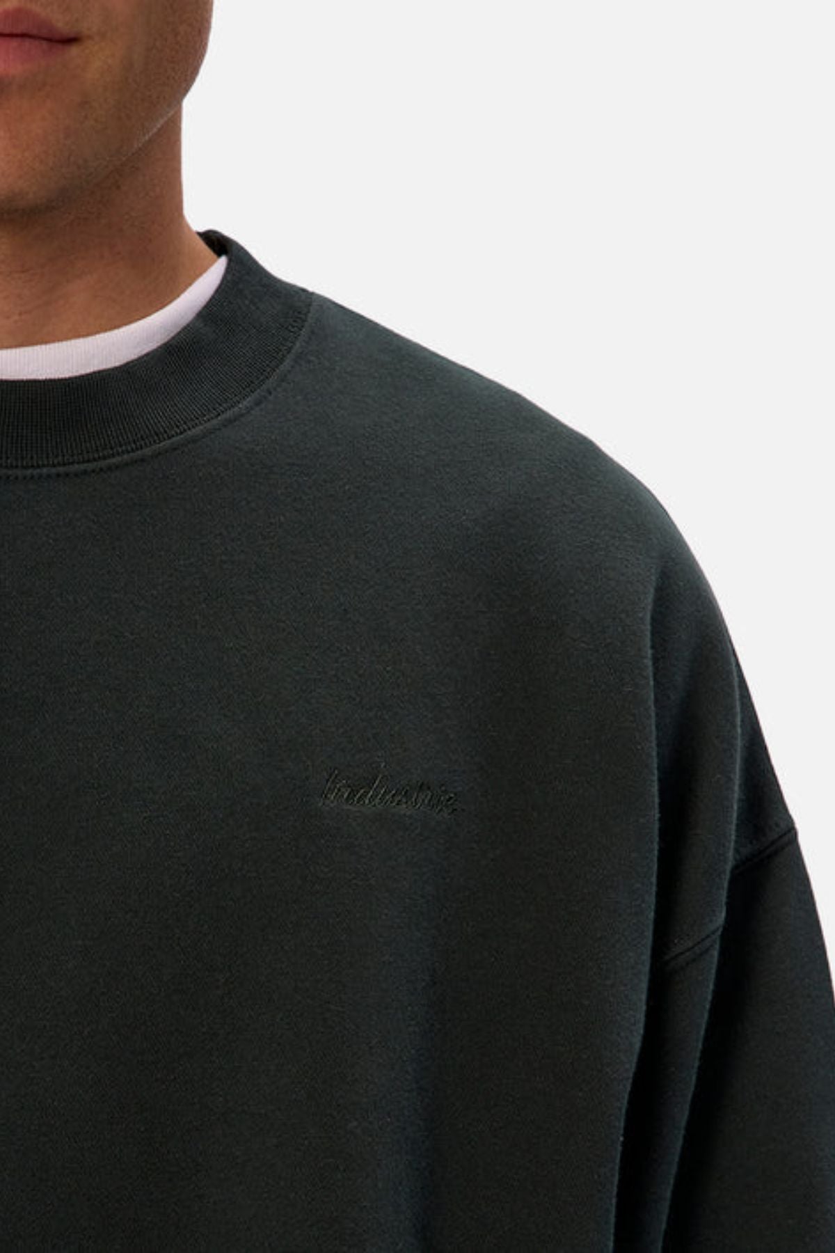A person wears an oversized, dark green Industrie The New Del Sur Washed Sweat with subtle chest embroidery, layered over a white T-shirt. Only the lower face and upper torso are shown in the photo.