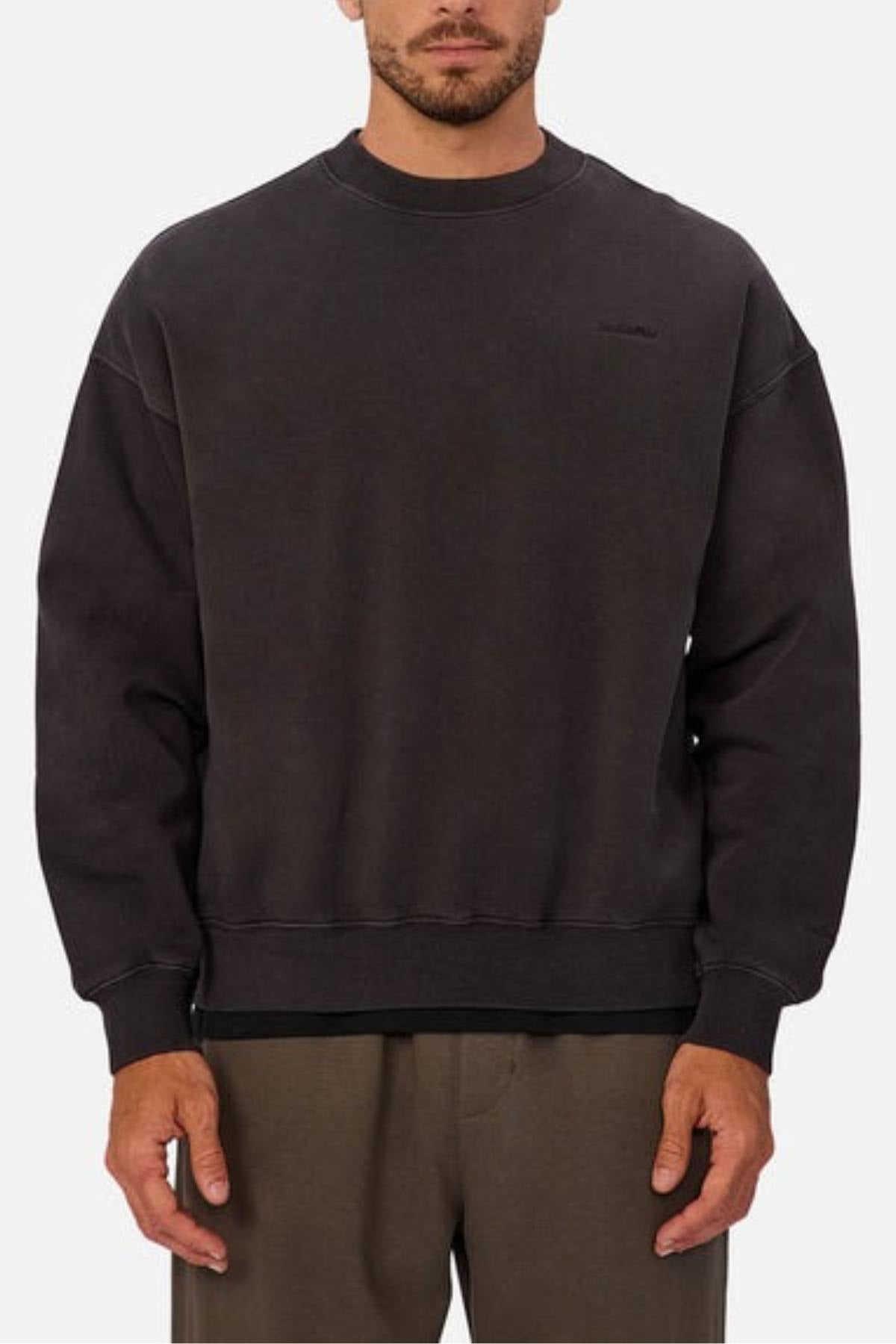 A man wears the Industrie The New Del Sur Washed Sweat, an oversized fit black crew neck sweatshirt with long sleeves and minimal detailing, paired with brown pants, standing against a plain light background.