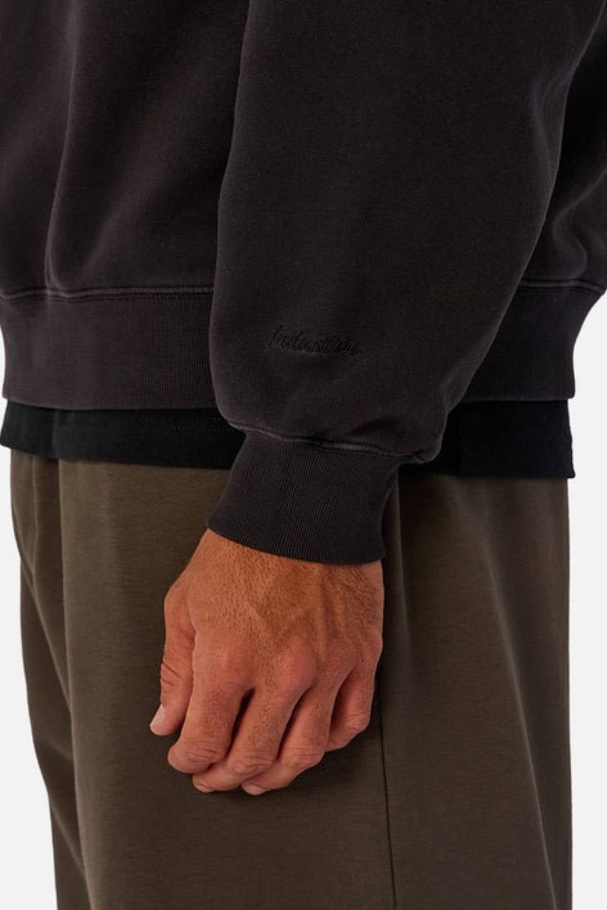 A person wears the Industrie The New Del Sur Washed Sweat in a dark, oversized fit with brown pants, their arm relaxed by their side. Only the lower torso and hand are visible, with the sleeve subtly showing embroidered Industrie branding.