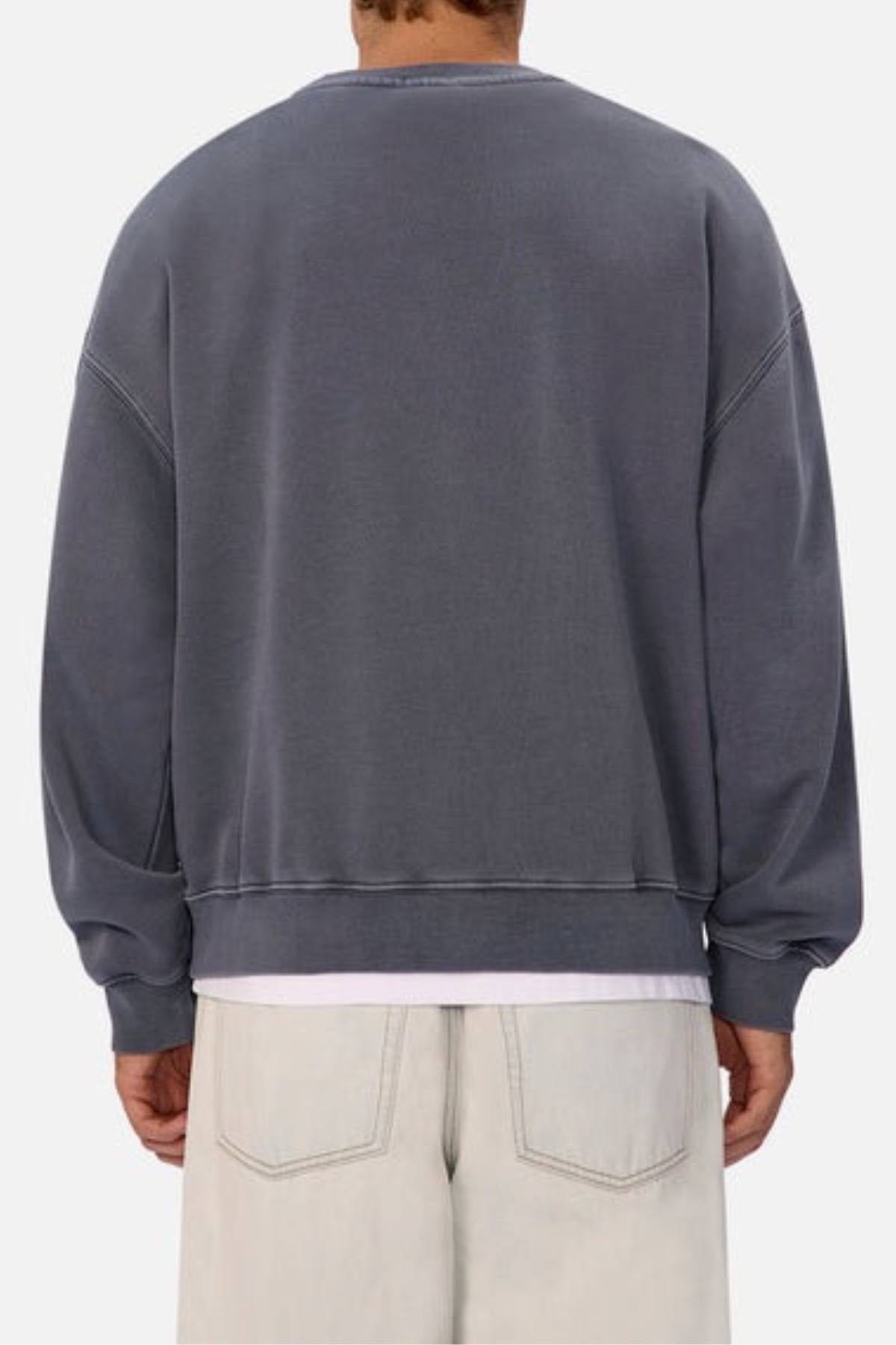 A person is seen from behind wearing an oversized Industrie The New Del Sur Washed Sweat in faded gray, layered over a white shirt and light pants, against a plain neutral background.