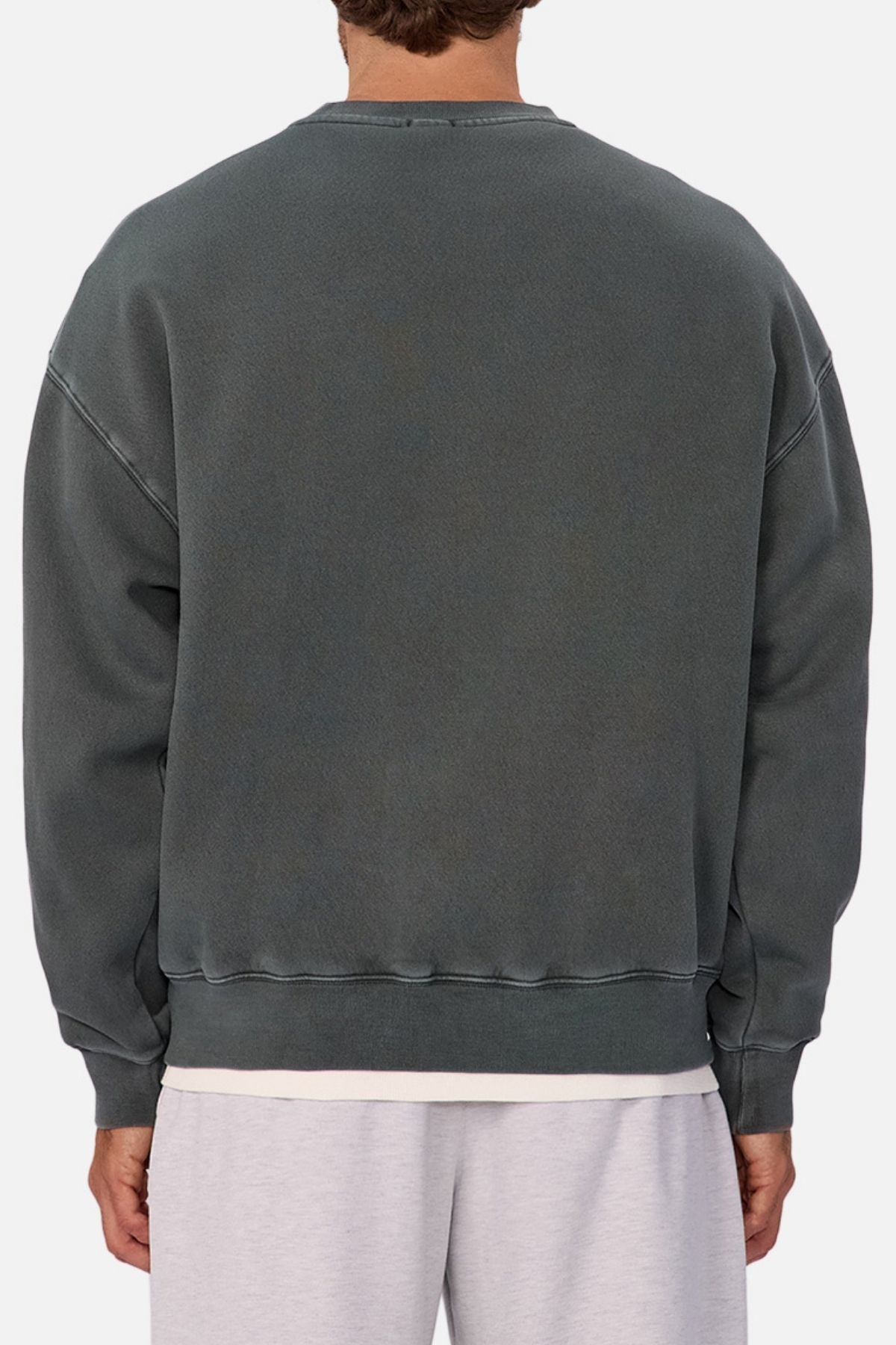 A person is seen from the back wearing an oversized Industrie The New Del Sur Washed Sweat in faded dark gray over a white T-shirt and light gray shorts. The focus is on the clothing, with a subtle Industrie logo and only part of the head and legs visible.