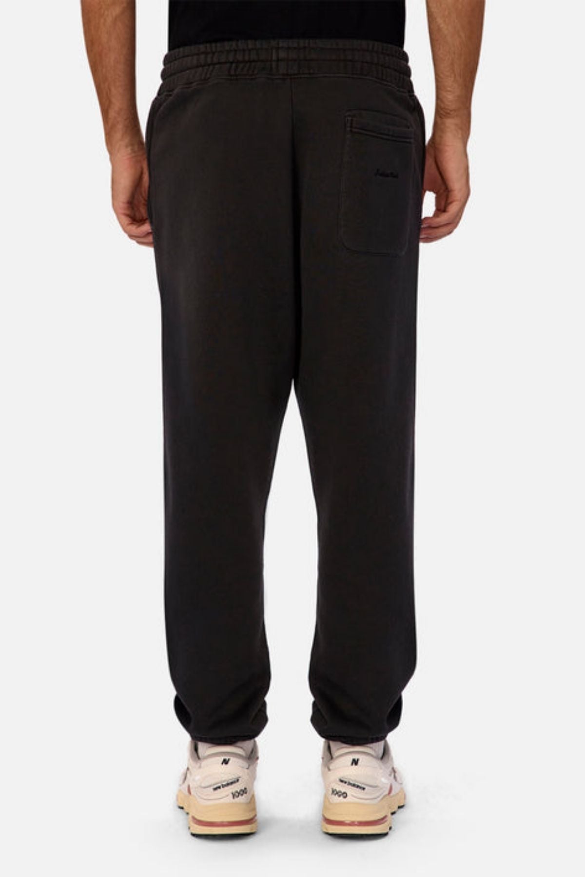 A person faces away, wearing Industrie The New Del Sur Washed Trackpant in black with a drawstring waist and elastic cuffs, paired with a black shirt and white New Balance sneakers against a plain, light background.