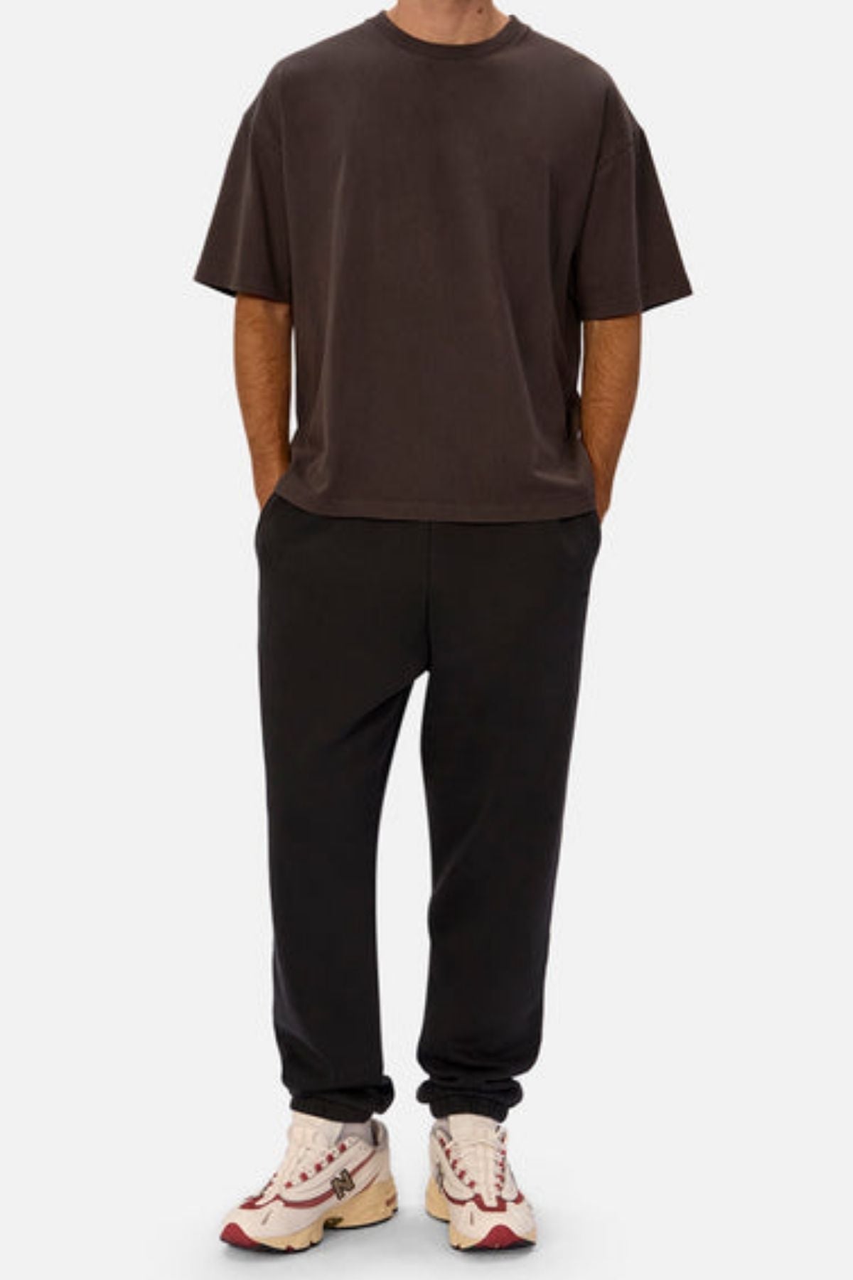 Wearing an oversized dark brown tee, Industrie The New Del Sur Washed Trackpant in black with elastic cuffs, and white sneakers with red and beige accents, a person stands hands-in-pockets against a plain background.